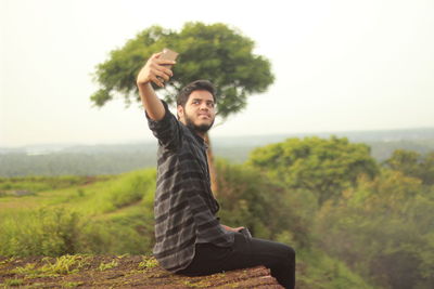 Young man taking selfie with smart phone while sitting on mountain against sky