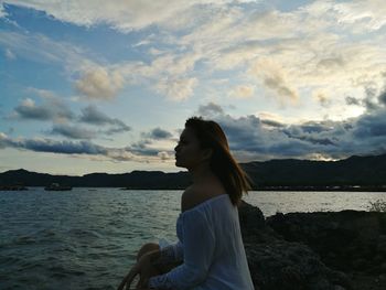 Side view of woman sitting by river against sky at dusk