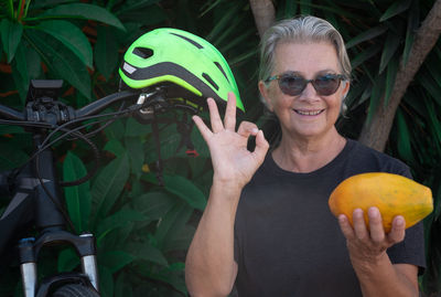 Portrait of smiling senior woman gesturing ok sign while holding papaya by bicycle and cycling helmet against plants