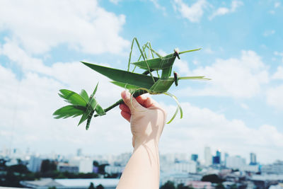 Cropped hand holding grasshoppers made from leaves against sky