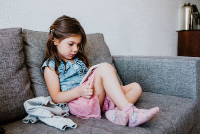 Full body of adorable ethnic little girl in casual clothes and socks relaxing on comfortable sofa and playing video game with smartphone