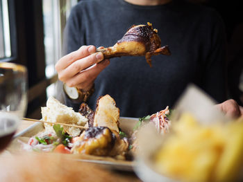 Crop unrecognizable man in casual wear eating grilled chicken leg while sitting at table with tray with assorted dishes in fast food restaurant