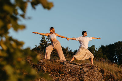 Young adult couple holding by hands, man and woman practicing yoga outdoors on the rock at sunset	
