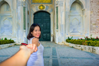 Portrait of smiling young woman holding hands while standing against built structure