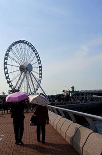 Rear view of women with umbrellas while walking by ferris wheel against sky