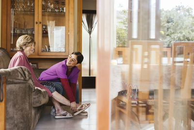 Happy caregiver helping senior woman to put on sandals in living room