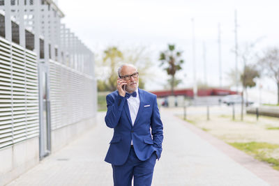 Confident senior man in suit talking through smart phone while walking on footpath against sky