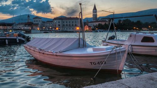 Sailboats moored on sea by city against sky during sunset