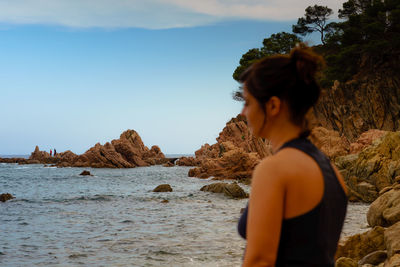 Rear view of woman standing on rock by sea against sky
