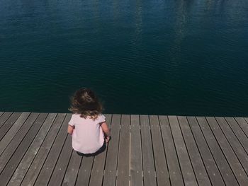 Rear view of girl sitting on jetty over lake during sunny day