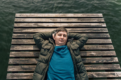 A man lies on a wooden plank pier on the lake in a jacket and jumper with his eyes closed