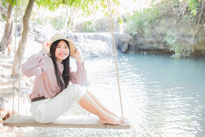 Portrait of smiling young woman sitting on swing over lake