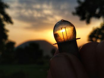 Close-up of hand holding lighting equipment against sky during sunset