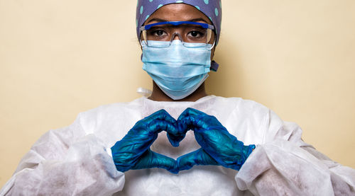 Portrait of young woman wearing mask making heart shape against colored background