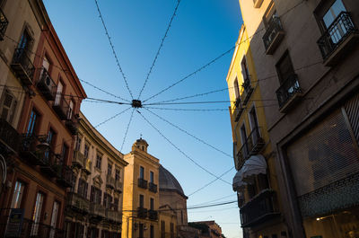 Low angle view of string lights hanging amidst buildings against blue sky