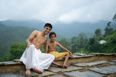 A young man sitting on the roof with his younger brother,both wearing dhoti, smiling into the camera