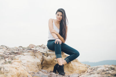Portrait of beautiful fashion model sitting on rock formation against clear sky