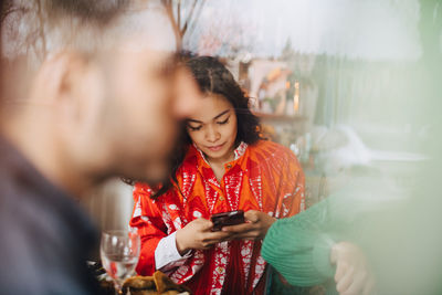 Young woman using smart phone while sitting with friends in restaurant seen through glass window