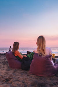 Rear view of woman sitting on beach against sky during sunset