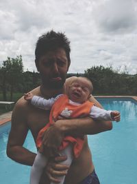 Portrait of man making face while holding crying baby girl standing by poolside against sky