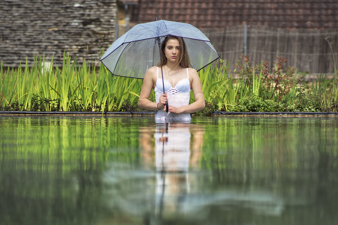 water, one person, young adult, real people, reflection, lake, women, young women, protection, leisure activity, umbrella, selective focus, adult, plant, day, waterfront, nature, outdoors, hairstyle, beautiful woman, rain