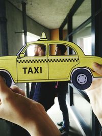 Cropped hand of man holding yellow taxi paper art against friends