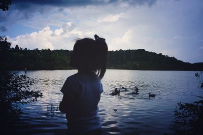 Rear view of silhouette girl standing by lake against sky