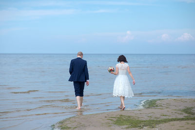 Rear view full length of bride and bridegroom walking at beach