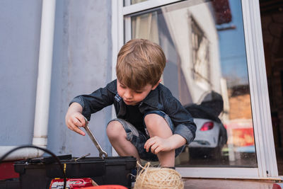 Curious kid taking wrench out of plastic container between glass door and twine ball in daytime
