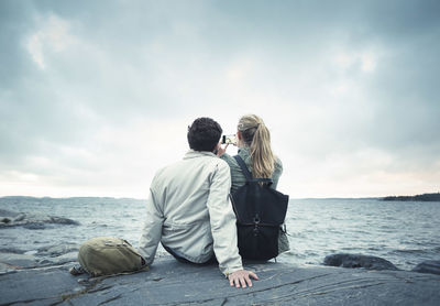 Rear view of couple taking selfie while sitting on rock by sea
