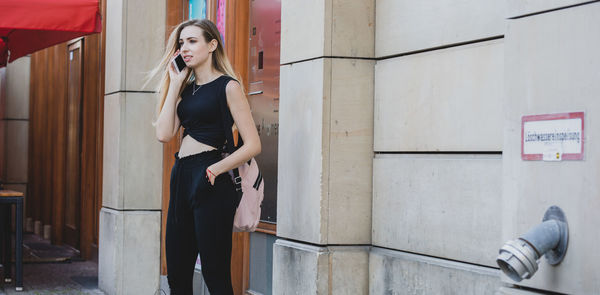 Beautiful young woman using smart phone while standing against wall