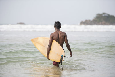 Shirtless man with surfboard wading in sea against clear sky