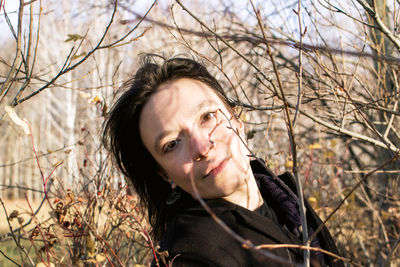 A young woman with long brown hair wearing a black coat stands in the middle of some bushes. 