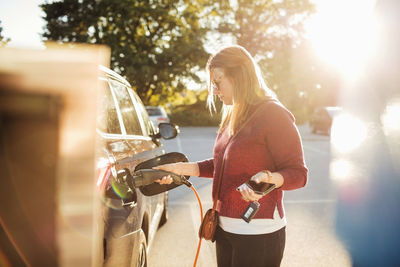 Woman holding smart phone while charging electric car at station on sunny day