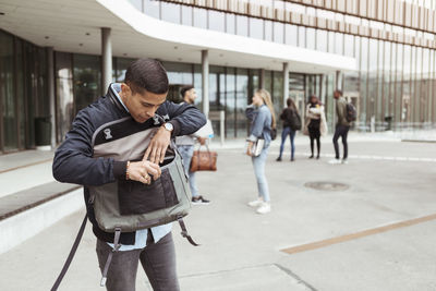 Young student searching in bag while friends standing in background at university