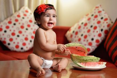 Full length of cheerful shirtless baby girl eating watermelon while sitting on table