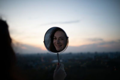 Reflection of smiling woman in mirror against sky during sunset