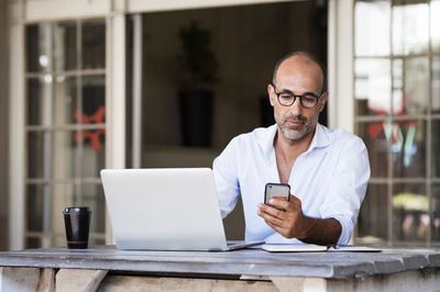 Businessman using smart phone while sitting with laptop computer at table in office