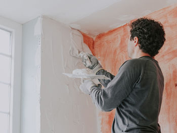 A young caucasian guy applies fresh putty with a small spatula on the wall near the window