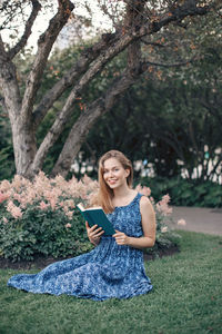 Portrait of smiling young holding book while woman sitting on grass against trees