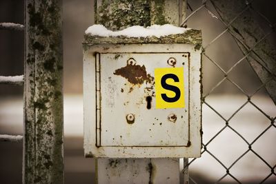 Close-up of alphabet s on fuse box attached to chainlink fence