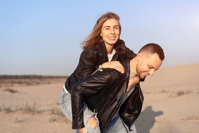 Boyfriend carrying girlfriend on back on sand dune against sky