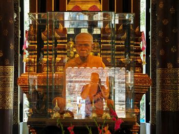 Reflection of man photographing monk sculpture on glass at temple