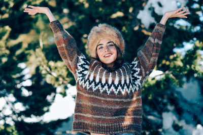 Portrait of smiling young woman with arms outstretched standing against trees during winter