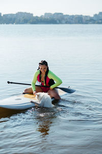 Dog japanese spitz swimming in the lake water and trying to get on the sup board with woman on it