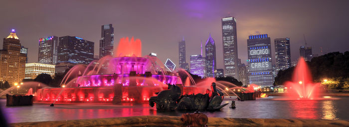 Illuminated modern buildings by river against sky at night