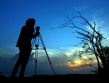 Silhouette of man standing with tripod against sky during sunset