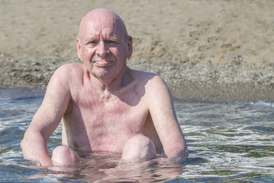 Old bald man sits in water sea and sunbathes in sun. sunny autumn day. front view.