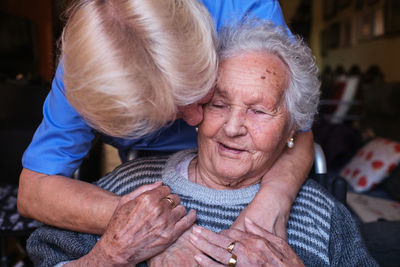 Caring nurse in blue uniform embracing and kissing elderly disabled woman with gray hair sitting in wheelchair in light room at home