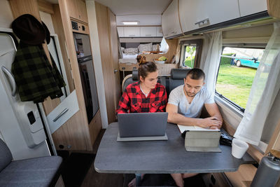 Smiling traveling couple of freelancers using laptop and tablet while working on project remotely at table in caravan